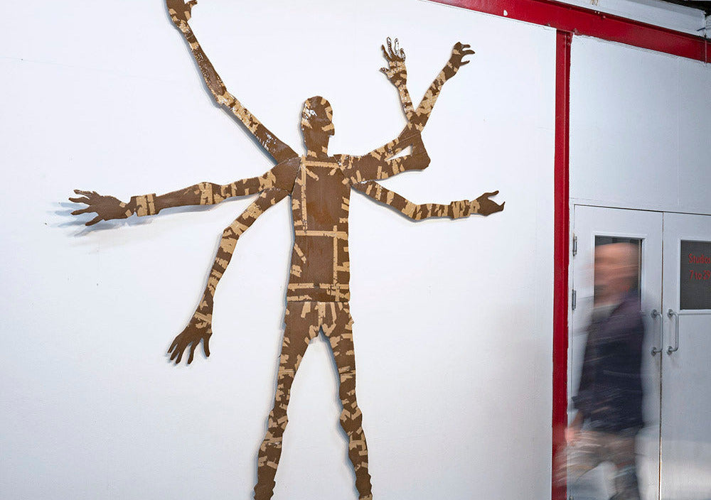 Artistic depiction of a multi-armed figure on a white wall with a person walking by.